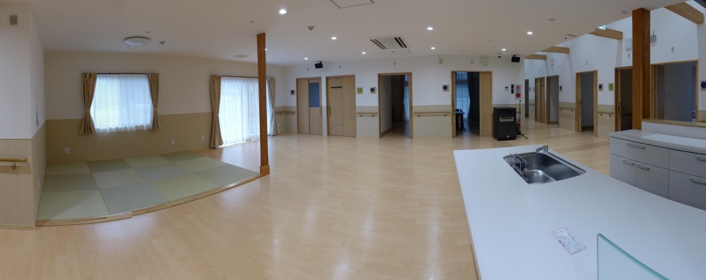 介護施設の広々とした共用スペース。明るく清潔な空間で複数の出入口と設備が配置されている。